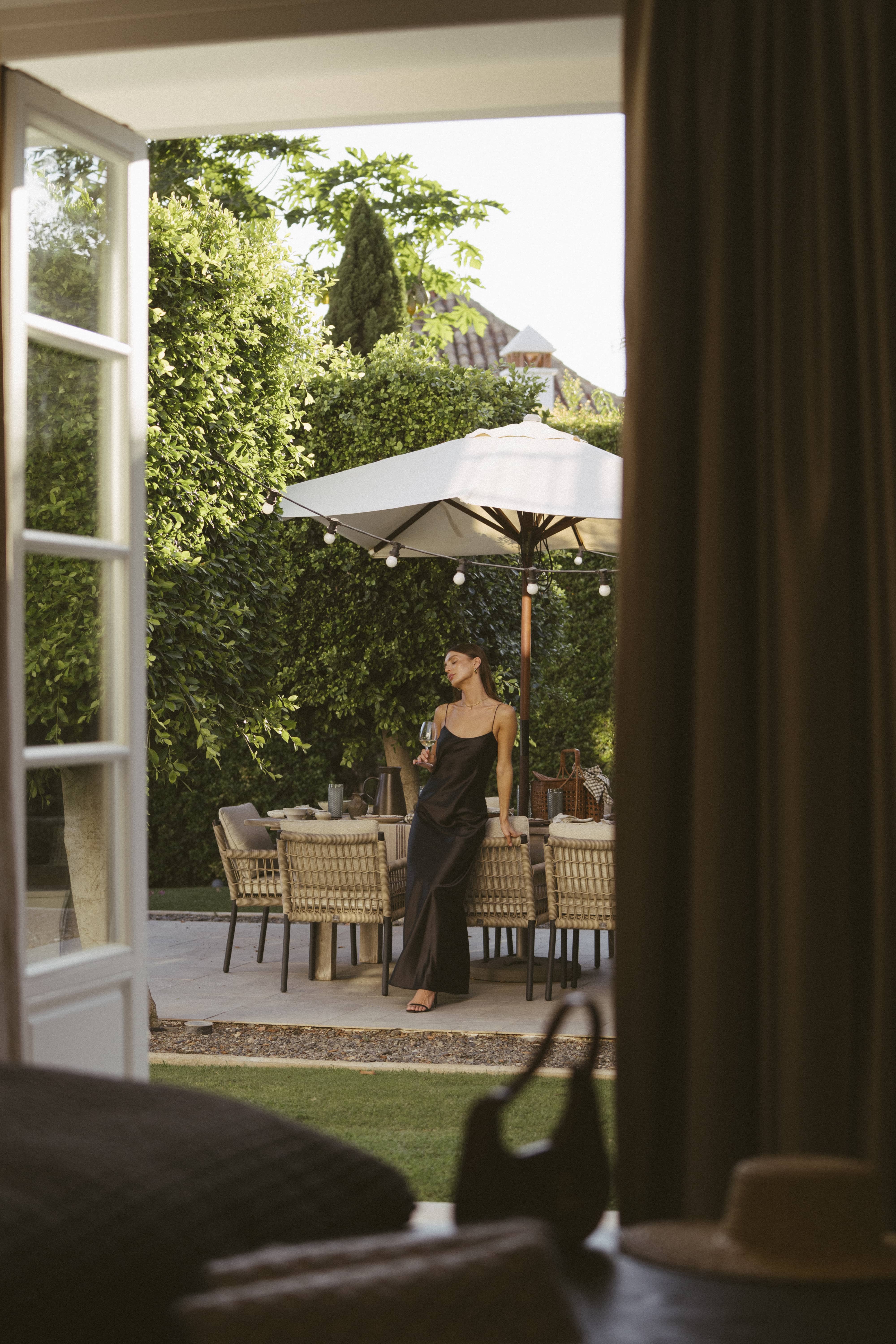 A woman wearing a black dress, standing under an umbrella in a garden near a table with chairs. The scene is framed by open doors with a curtain and lush greenery in the background.